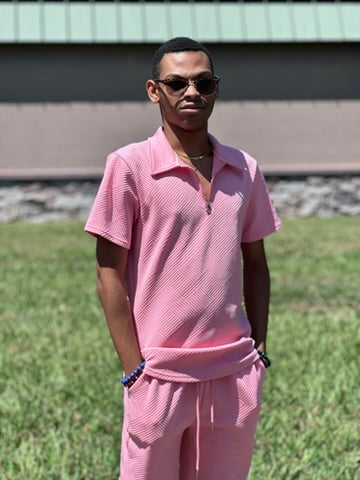 Jay is wearing a matching light pink textured polo shirt and shorts set, styled with dark sunglasses and a gold chain necklace, standing outdoors on a grassy lawn.