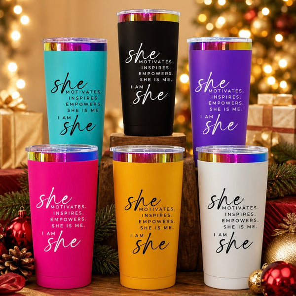 Six insulated stainless steel tumblers with clear lids and metallic rims, each in a different color (teal, black, purple, pink, yellow, white), arranged on a wooden surface with holiday lights and ornaments in the background. Each tumbler has the phrase 'She Motivates.'