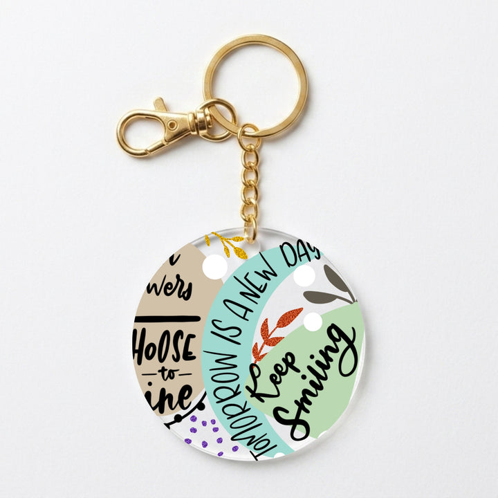 A round acrylic keychain charm with a gold-tone clasp and chain, featuring a pastel multicolor design with text including 'Choose to shine' and 'Tomorrow is a new day' on a light gray background.