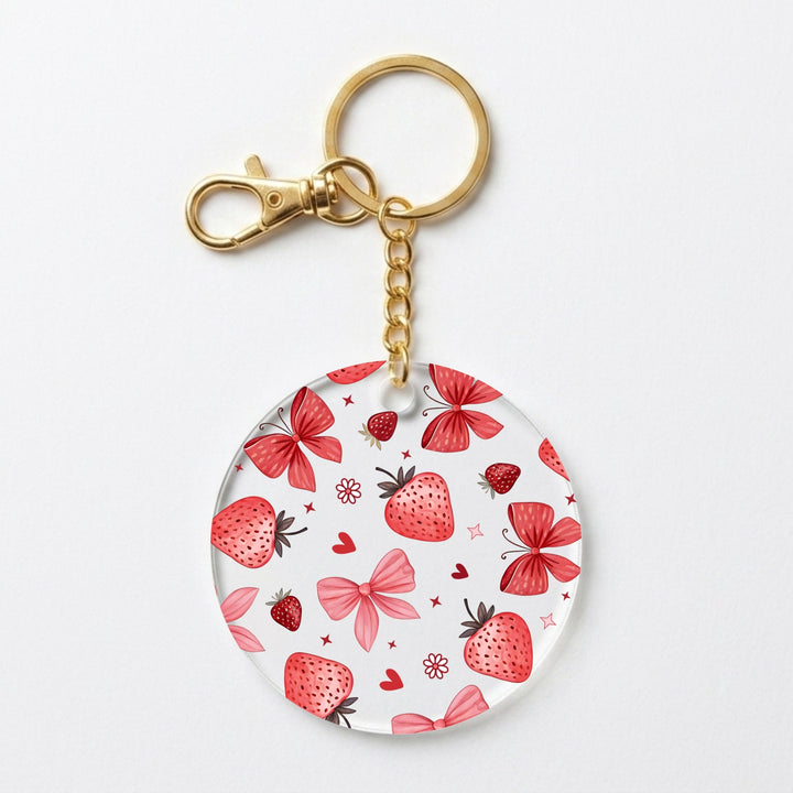 A round acrylic keychain charm featuring a pattern of red strawberries, pink bows, and butterfly bows on a white background, attached to a gold-tone split ring and lobster clasp.