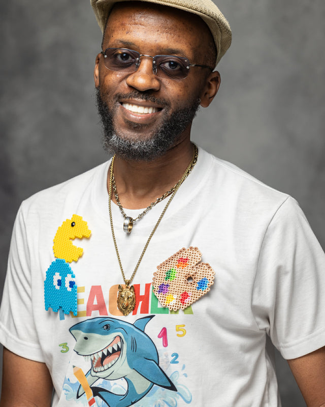 William is smiling and wearing a beige cap, sunglasses, and a white graphic t-shirt with a colorful dinosaur, shark, and paint palette design, set against a textured gray studio background.