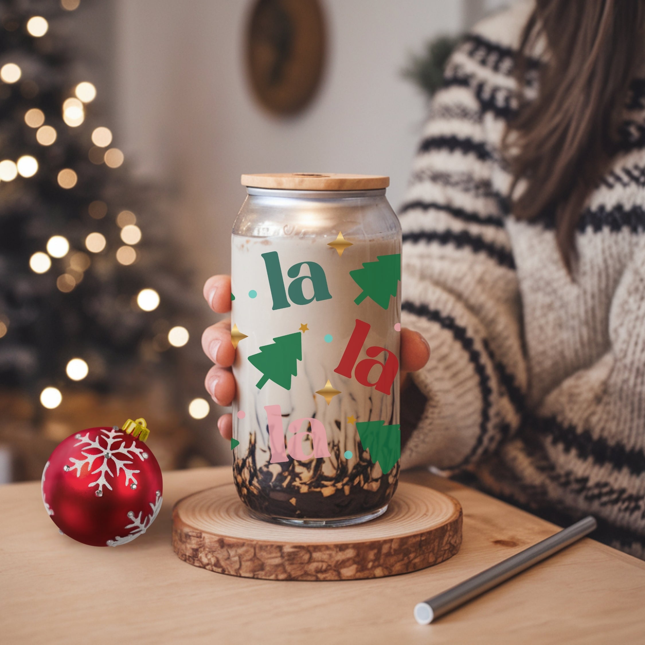 Person holding a festive-themed glass with Christmas decorations and lights, blurred Christmas tree in the background.