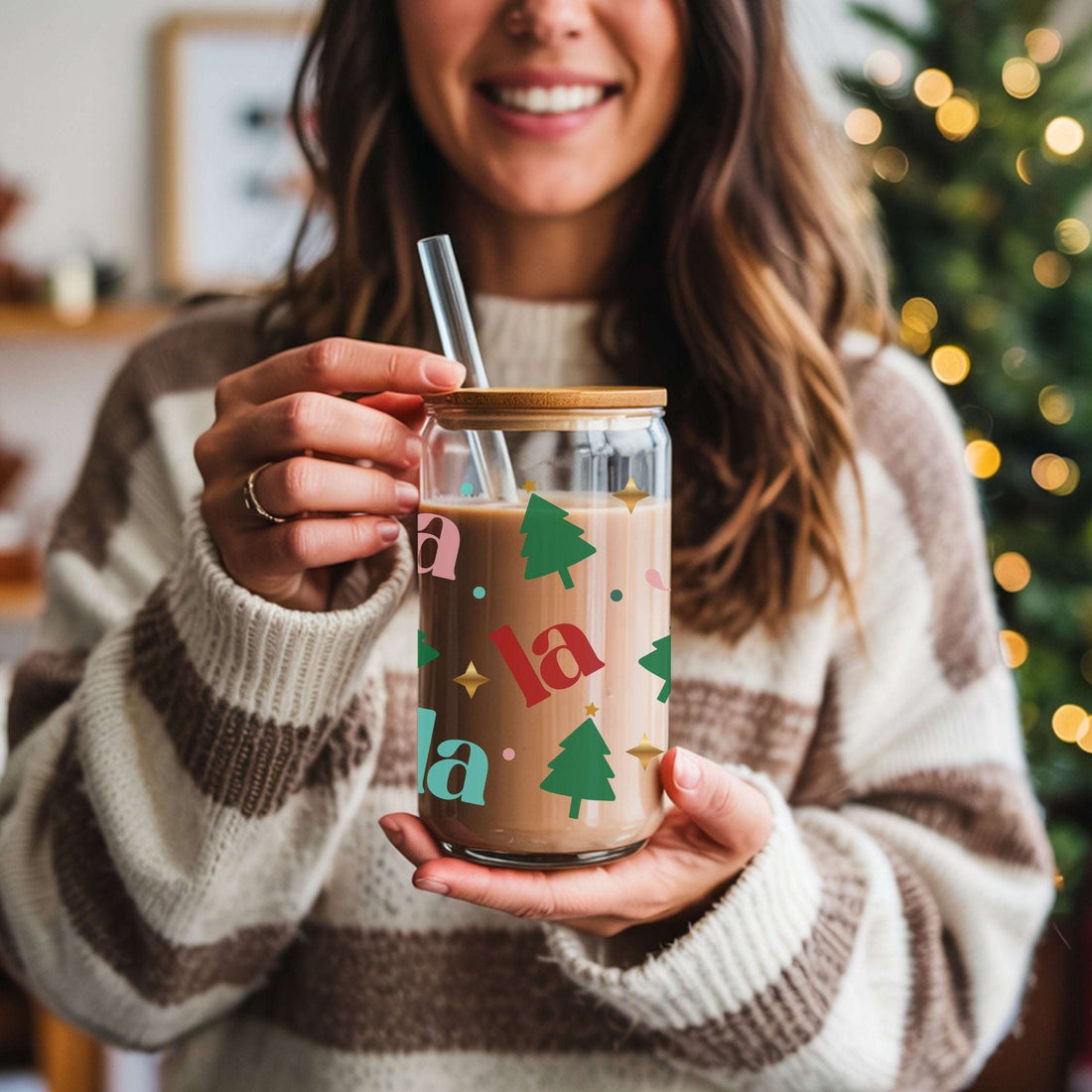 Woman holding a glass mug with Christmas-themed design and straw, blurred Christmas tree in the background