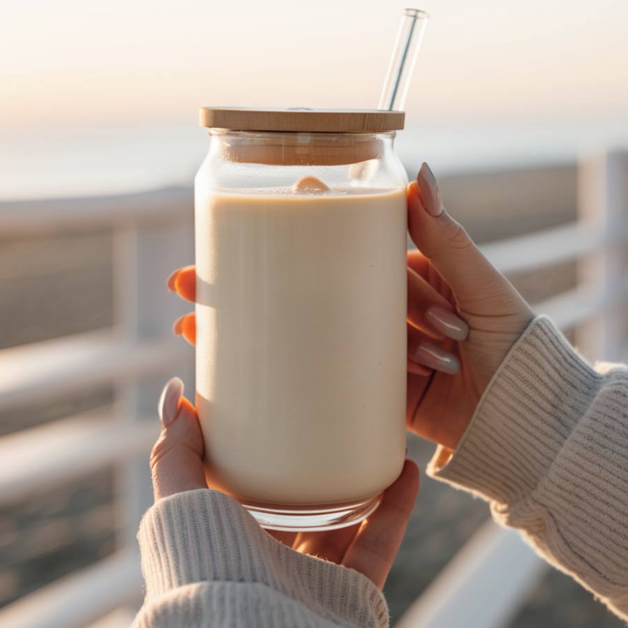 Person holding a 16oz can glass cup with a wooden lid and straw filled with a creamy liquid, outdoors.