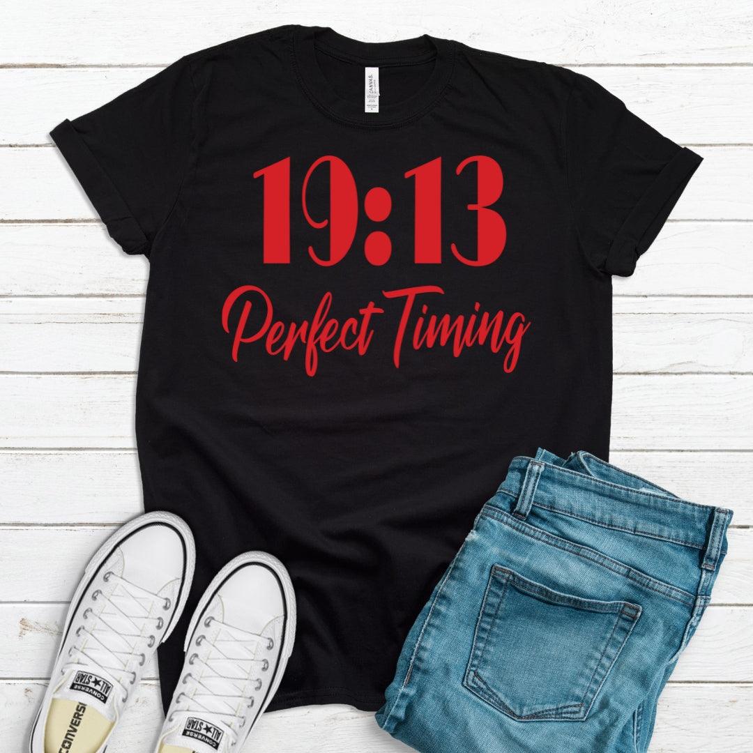 Black t-shirt with red text '19:13 Perfect Timing', white sneakers, and blue jeans on a wooden surface.