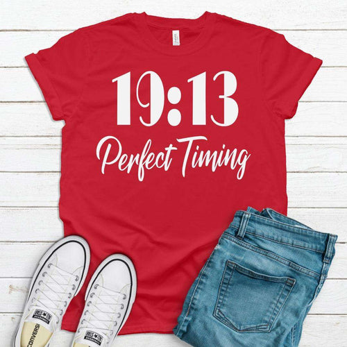 Red t-shirt with '19:13 Perfect Timing' text, white sneakers, and blue jeans on a wooden surface.