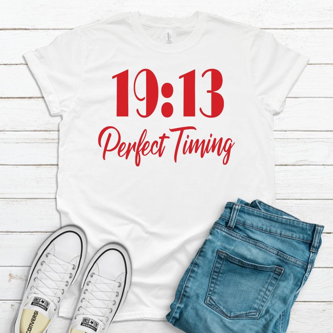 White t-shirt with '19:13 Perfect Timing' text, paired with jeans and sneakers on a wooden surface.