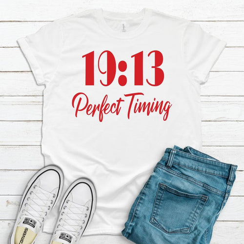 White t-shirt with '19:13 Perfect Timing' text, paired with jeans and sneakers on a wooden surface.