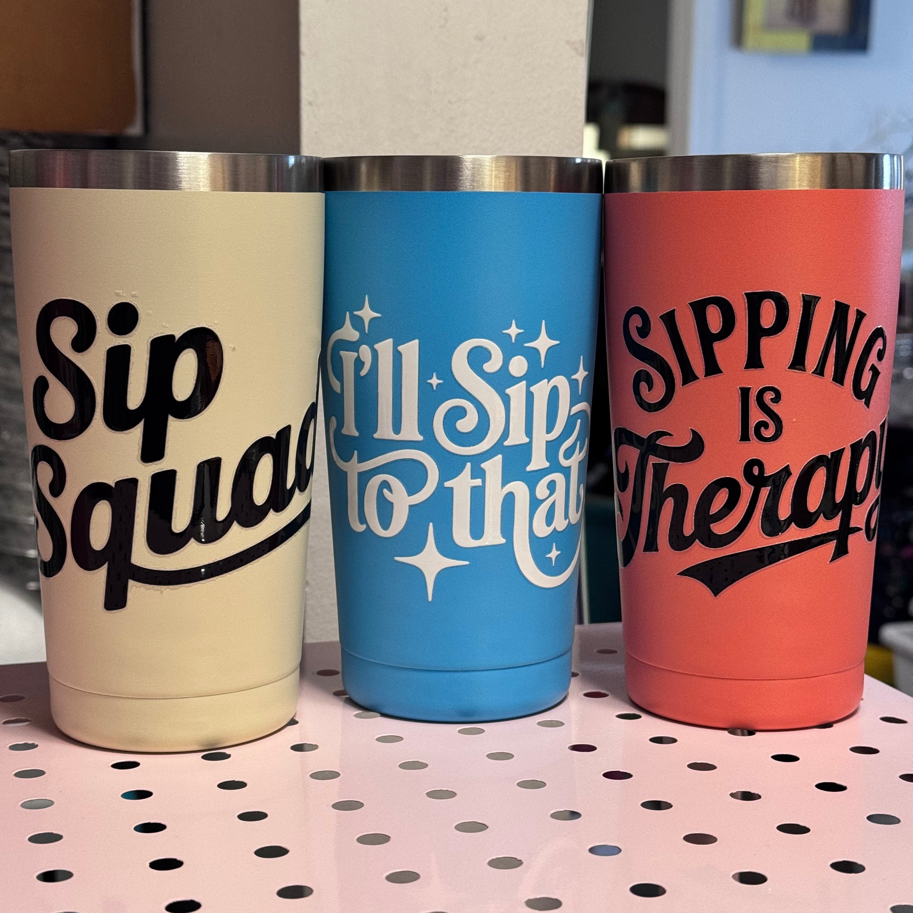 Three colorful tumblers with humorous text designs on a pegboard.