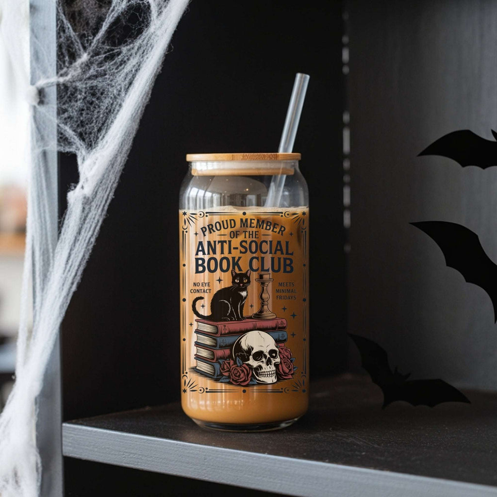 A Halloween-themed glass can cooler with a straw, filled with iced coffee and decorated with a gothic illustration of a black cat, books, skull, and roses. The text on the label reads 'PROUD MEMBER OF THE ANTI-SOCIAL BOOK CLUB'.