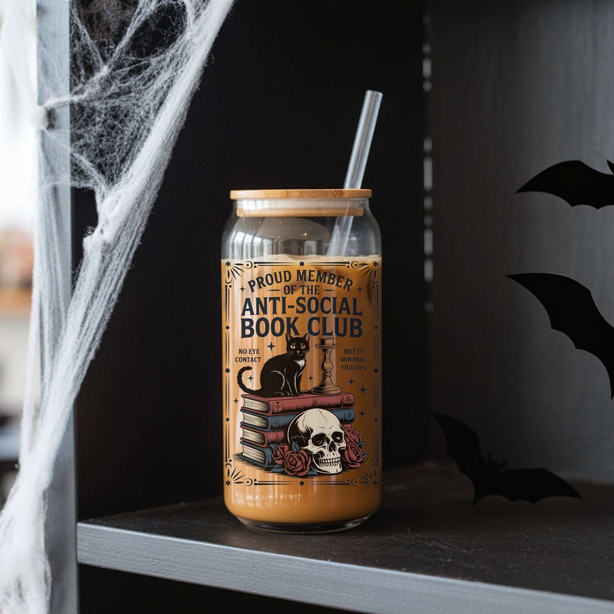 A Halloween-themed glass can cooler with a straw, filled with iced coffee and decorated with a gothic illustration of a black cat, books, skull, and roses. The text on the label reads 'PROUD MEMBER OF THE ANTI-SOCIAL BOOK CLUB'.