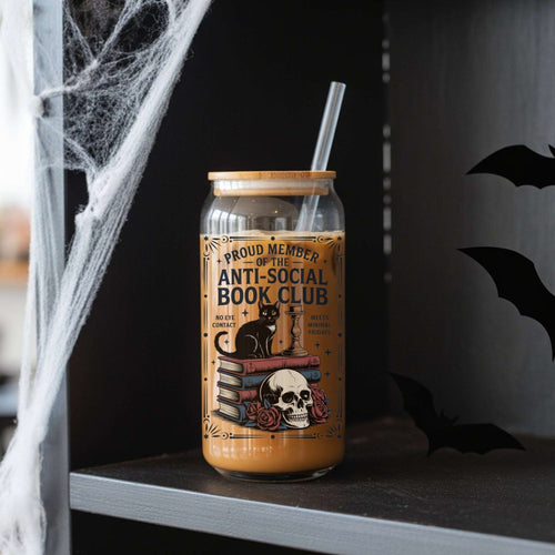 A Halloween-themed glass can cooler with a straw, filled with iced coffee and decorated with a gothic illustration of a black cat, books, skull, and roses. The text on the label reads 'PROUD MEMBER OF THE ANTI-SOCIAL BOOK CLUB'.