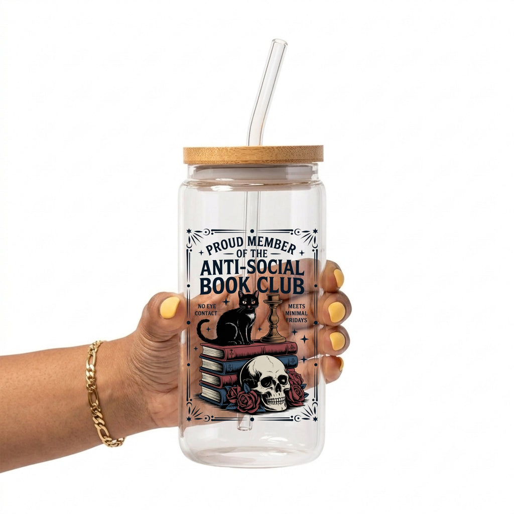 A hand with yellow nail polish holds a clear insulated tumbler with a bamboo lid and clear straw. The tumbler features a graphic design with a black cat, books, a skull, and roses, with the text 'PROUD MEMBER OF THE ANTI-SOCIAL BOOK CLUB'.