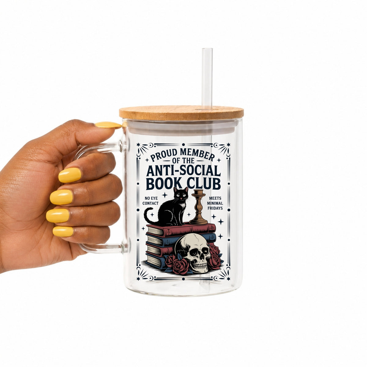 A hand with yellow nail polish holds a clear glass handled tumbler with a bamboo lid and clear straw. The tumbler features a graphic of a black cat sitting on a stack of books next to a skull and roses, with the text 'PROUD MEMBER OF THE ANTI-SOCIAL BOOK CLUB'.