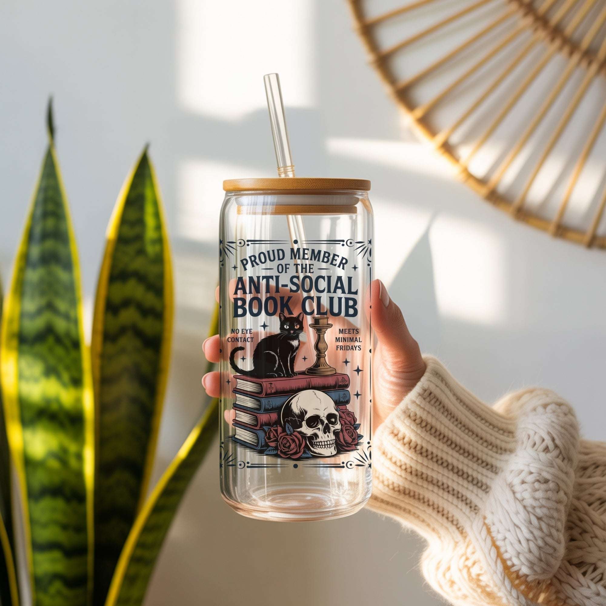 A hand wearing a cream knit sweater holds a clear glass can-style tumbler with a bamboo lid and straw. The tumbler has a decorative label reading 'Proud Member of the Anti-Social Book Club' with an illustration of a black cat, books, a skull, and roses.