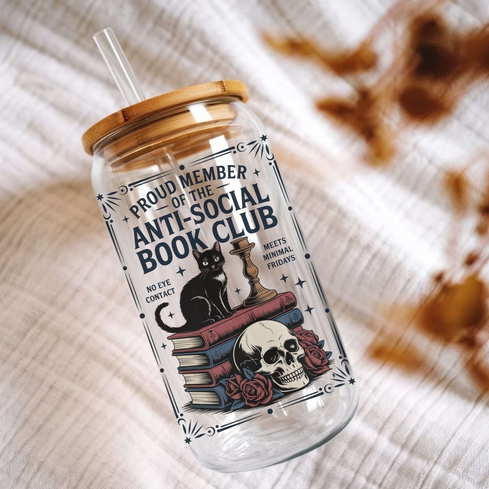 A clear glass tumbler with a bamboo lid and clear straw, printed with a gothic-style label that reads 'PROUD MEMBER OF THE ANTI-SOCIAL BOOK CLUB' with a black cat, chess piece, books, skull, and roses. The tumbler is photographed on a textured white fabric with blurred dried leaves in the background.