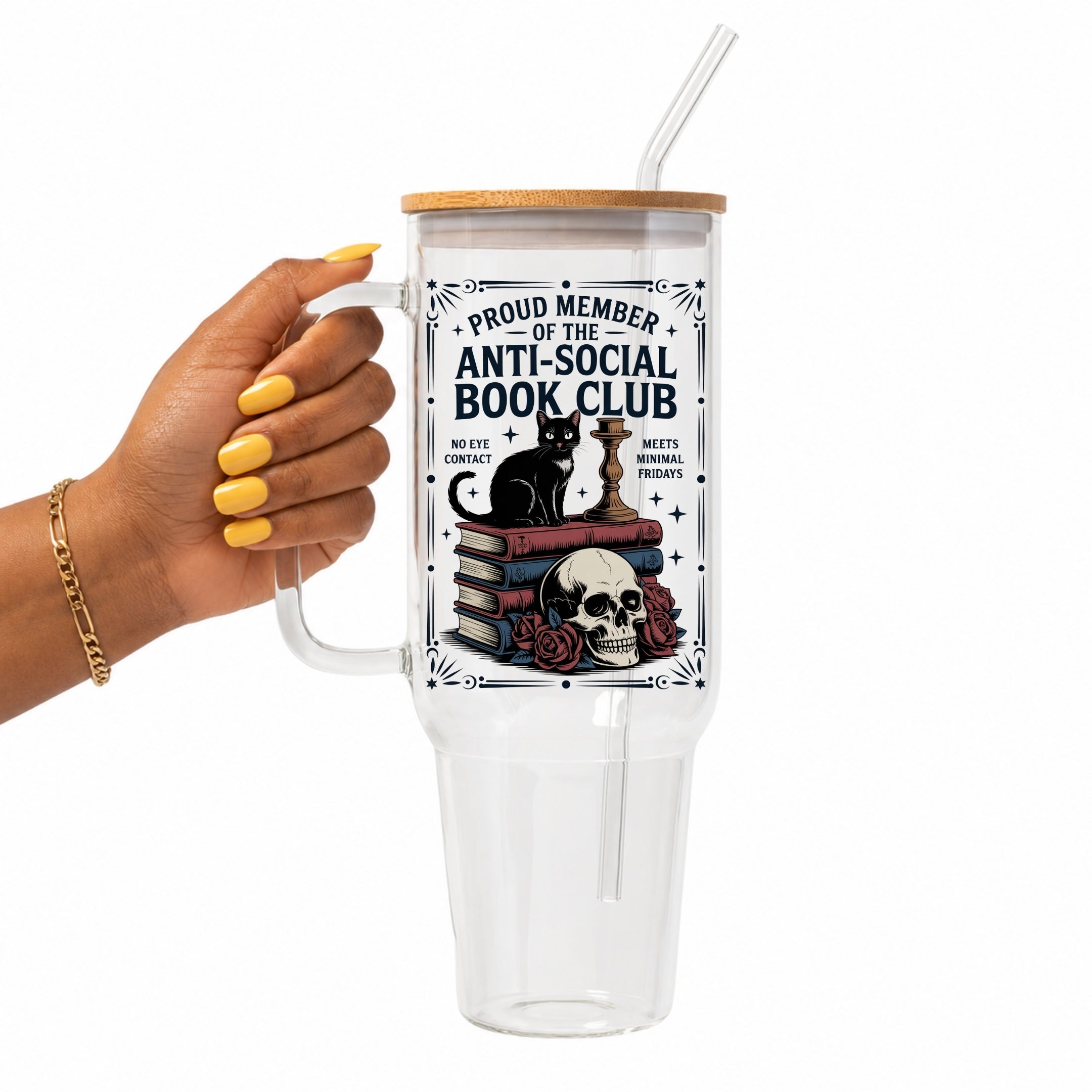 A hand with yellow nail polish holds a clear handled tumbler with a bamboo lid and clear straw. The tumbler features a graphic of a black cat on stacked books next to a skull and roses, with the text 'PROUD MEMBER OF THE ANTI-SOCIAL BOOK CLUB'.
