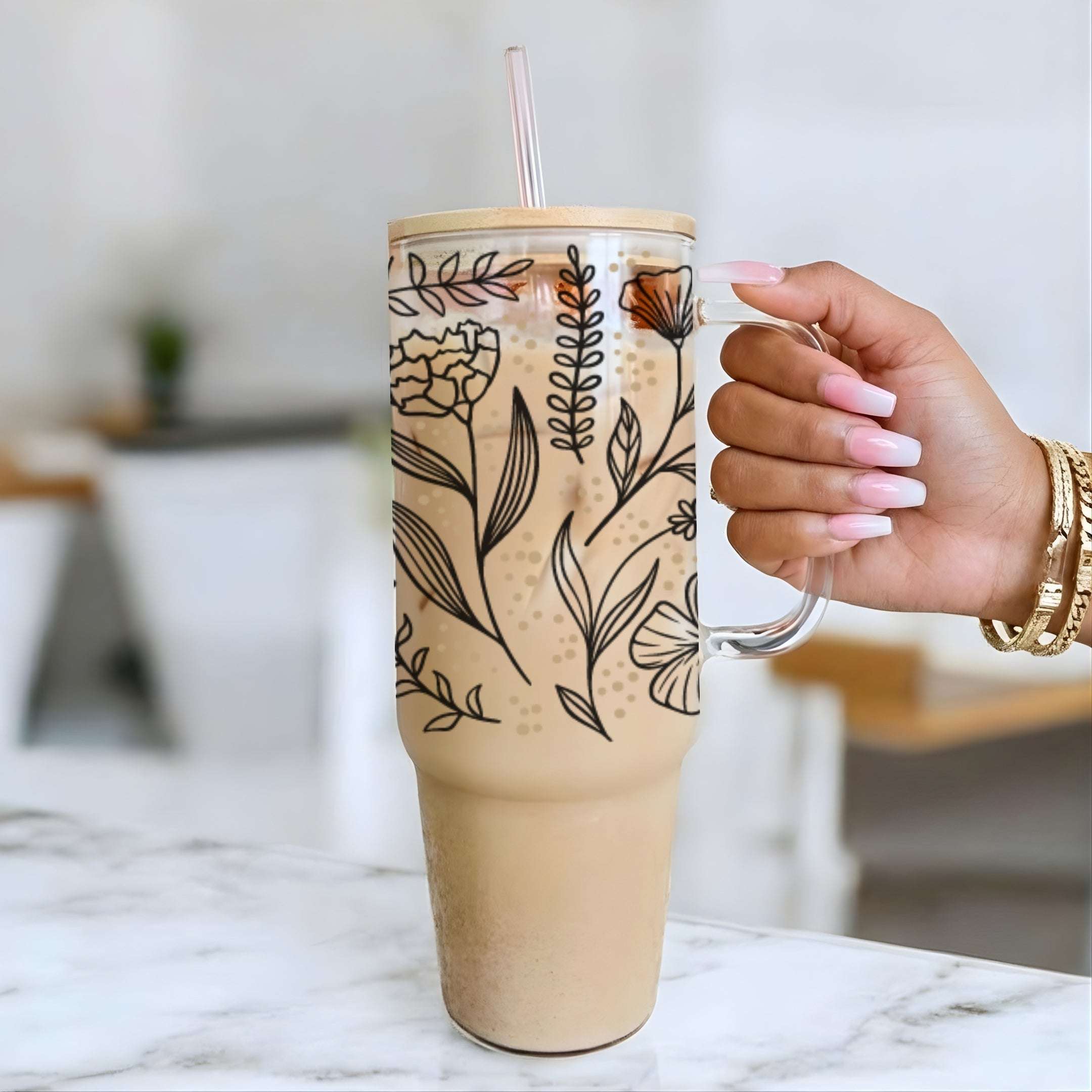 Nik's hand with pink and white manicured nails holds a tall insulated tumbler with a handle and clear straw, filled with an iced coffee drink. The tumbler has a black floral line-art pattern on a light beige background, set on a white marble countertop with a blurred indoor background.