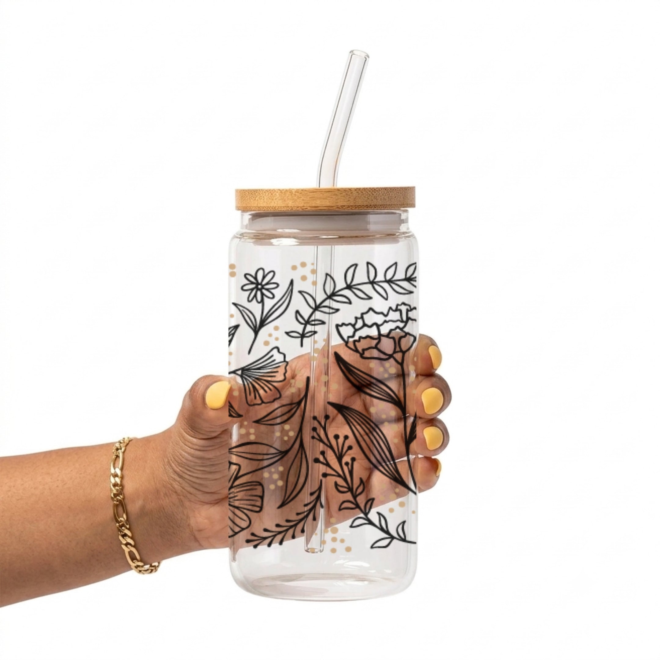 A hand holding a clear glass tumbler with a bamboo lid and clear glass straw, decorated with black botanical line art and small gold polka dots, against a white background.