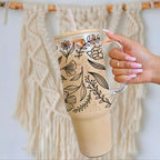 Nik's hand with pink manicured nails holds a tall clear glass tumbler with a handle, a bamboo lid, and a clear straw. The tumbler is decorated with black botanical line art and contains a light tan iced coffee.