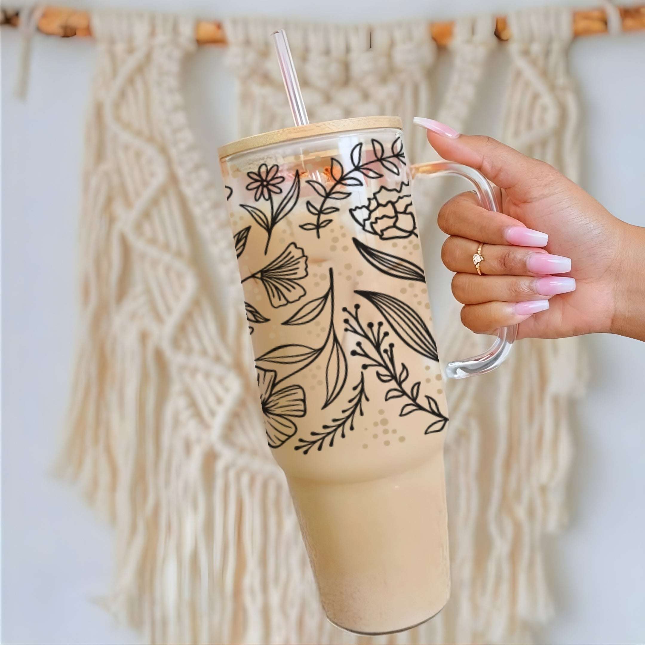 Nik's hand with pink manicured nails holds a tall clear glass tumbler with a handle, a bamboo lid, and a clear straw. The tumbler is decorated with black botanical line art and contains a light tan iced coffee.