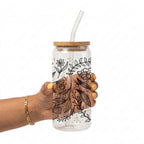 A hand with yellow nail polish holds a clear glass water bottle with a bamboo lid and a clear bent glass straw. The bottle is decorated with black botanical line art and small gold polka dots, set against a plain white background.