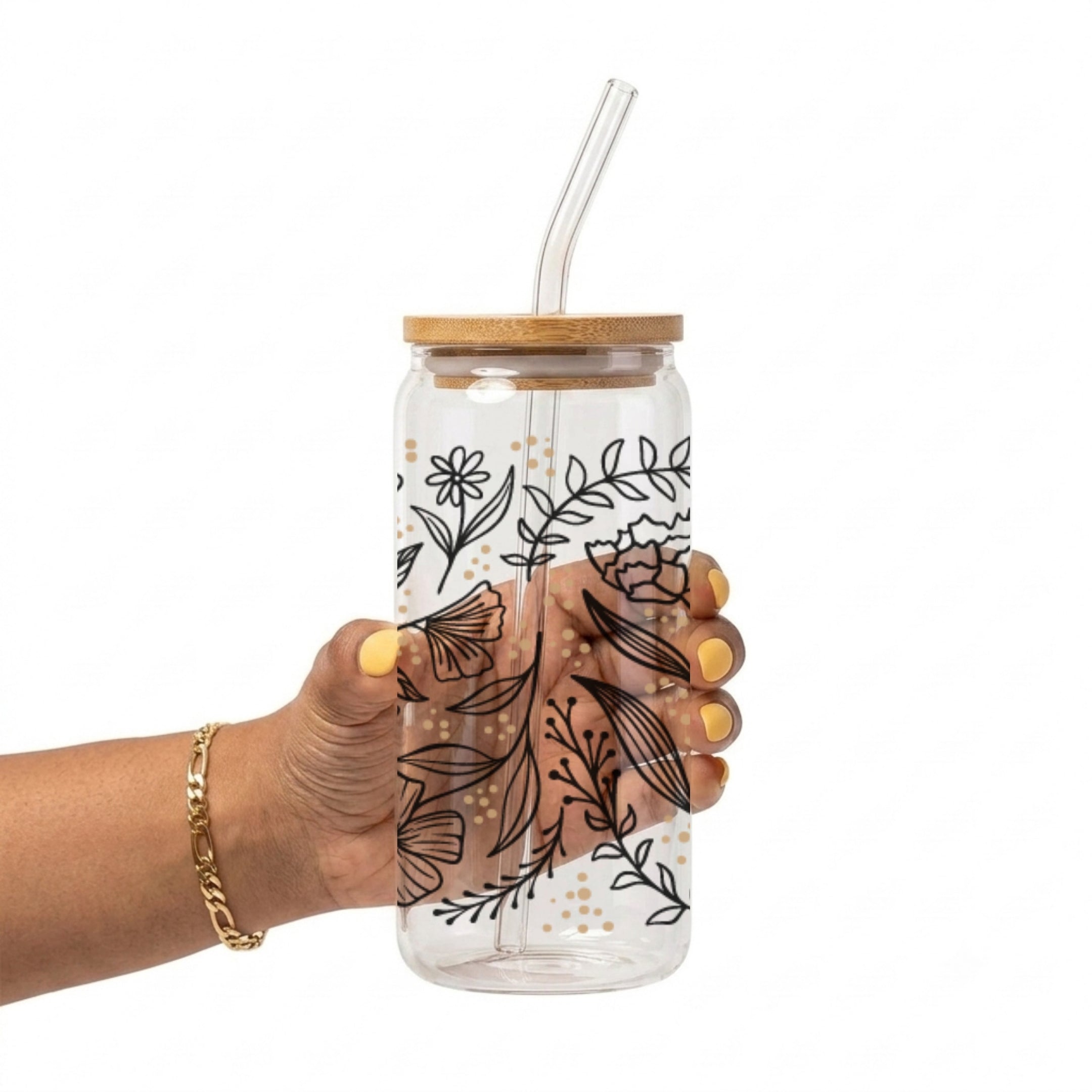 A hand with yellow nail polish holds a clear glass water bottle with a bamboo lid and a clear bent glass straw. The bottle is decorated with black botanical line art and small gold polka dots, set against a plain white background.