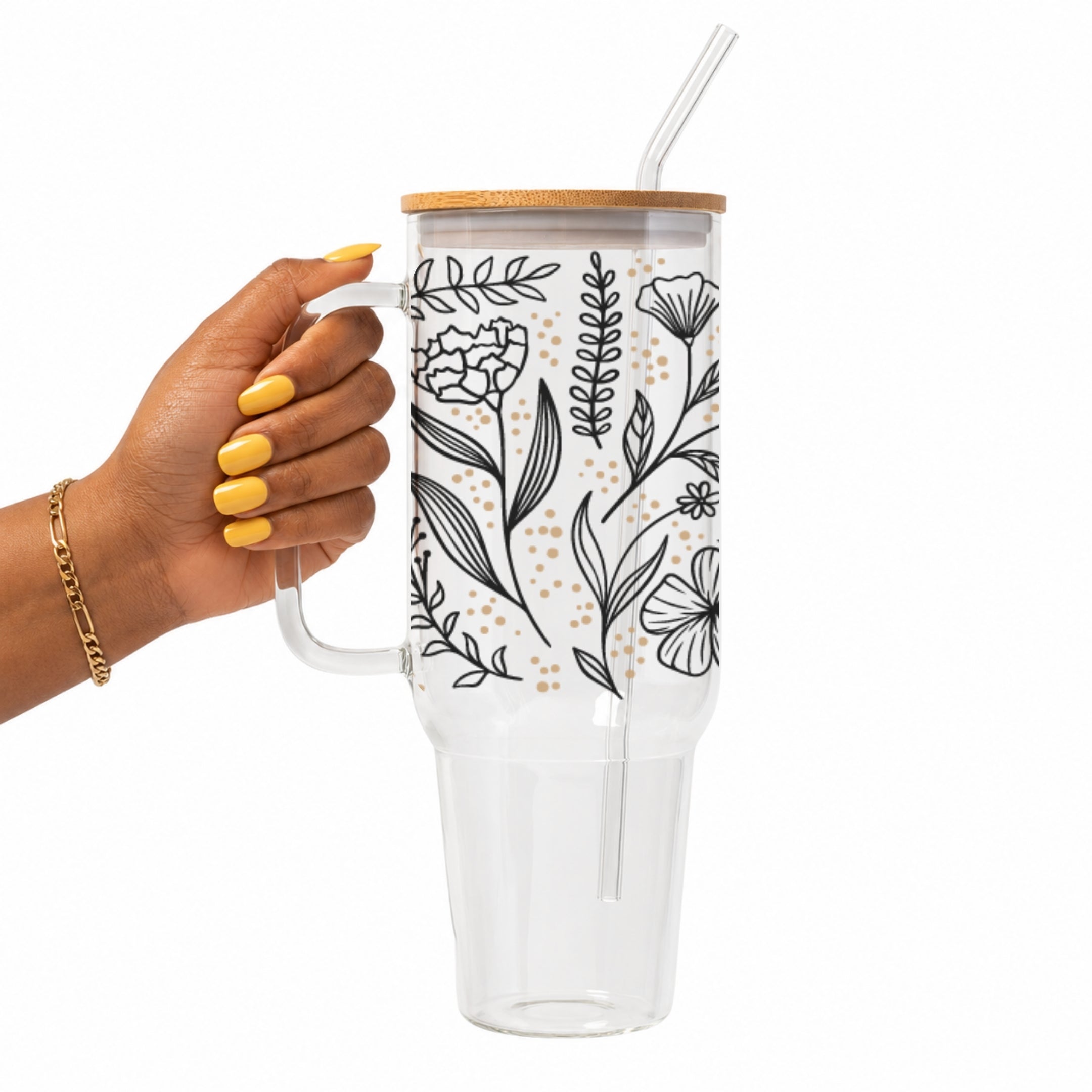 A hand with yellow nails holds a clear handled tumbler with a bamboo lid and clear straw. The tumbler has a white background decorated with black botanical line drawings and small gold polka dots.