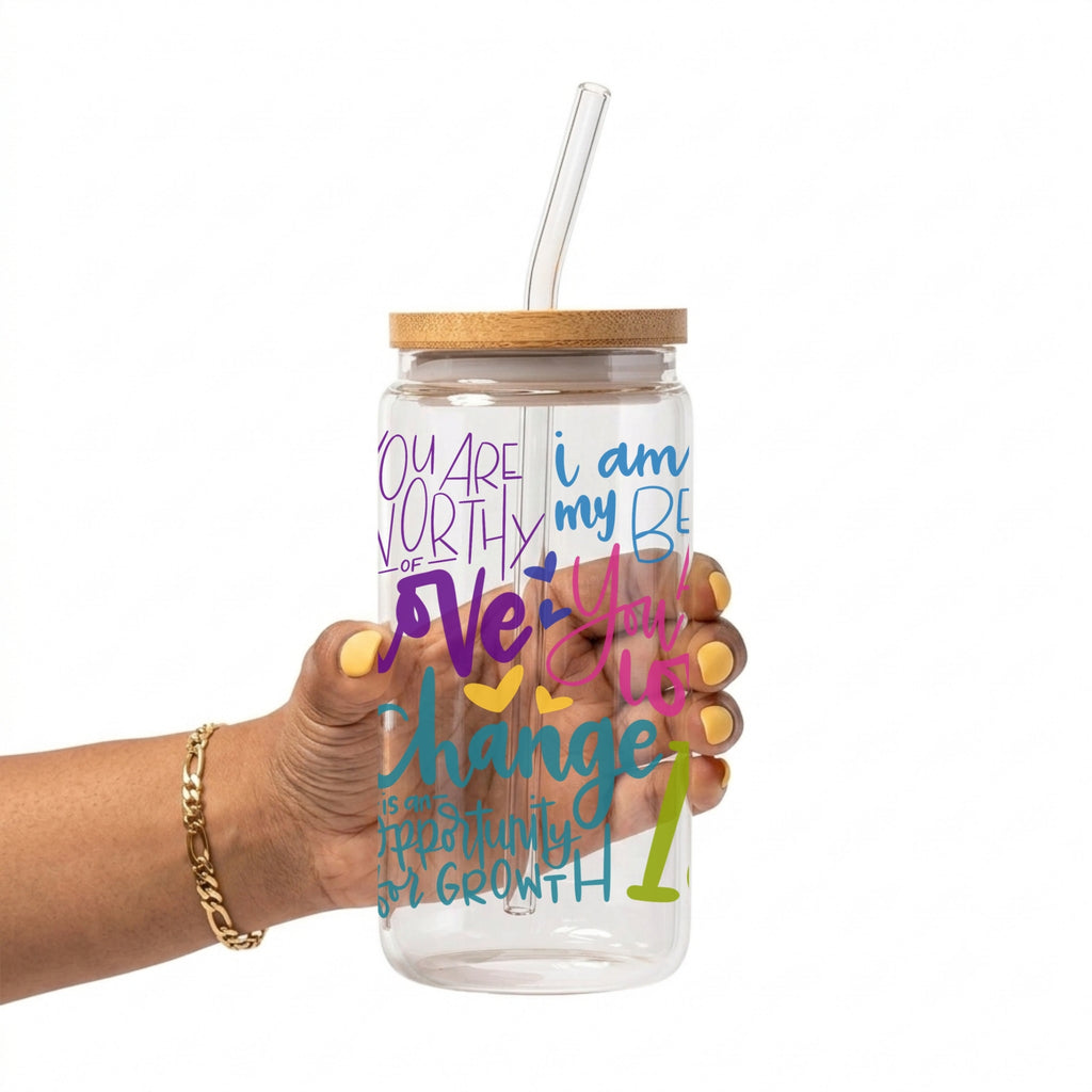 A hand holding a clear glass water bottle with a bamboo lid and clear straw. The bottle features colorful text in purple, blue, pink, yellow, and green.