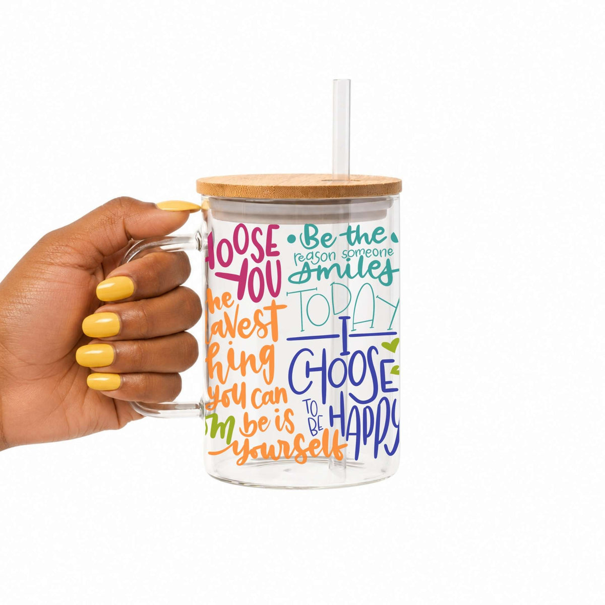 A hand with yellow nail polish holds a clear glass mug with a bamboo lid and straw. The mug features colorful hand-lettered positive affirmations on a white background.