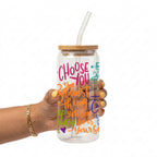 A hand holding a clear glass water bottle with a bamboo lid and clear glass straw. The bottle features colorful hand-lettered text that reads 'CHOOSE YOU 2 the bravest thing you can many be is yourself' in pink, orange, green, and blue.