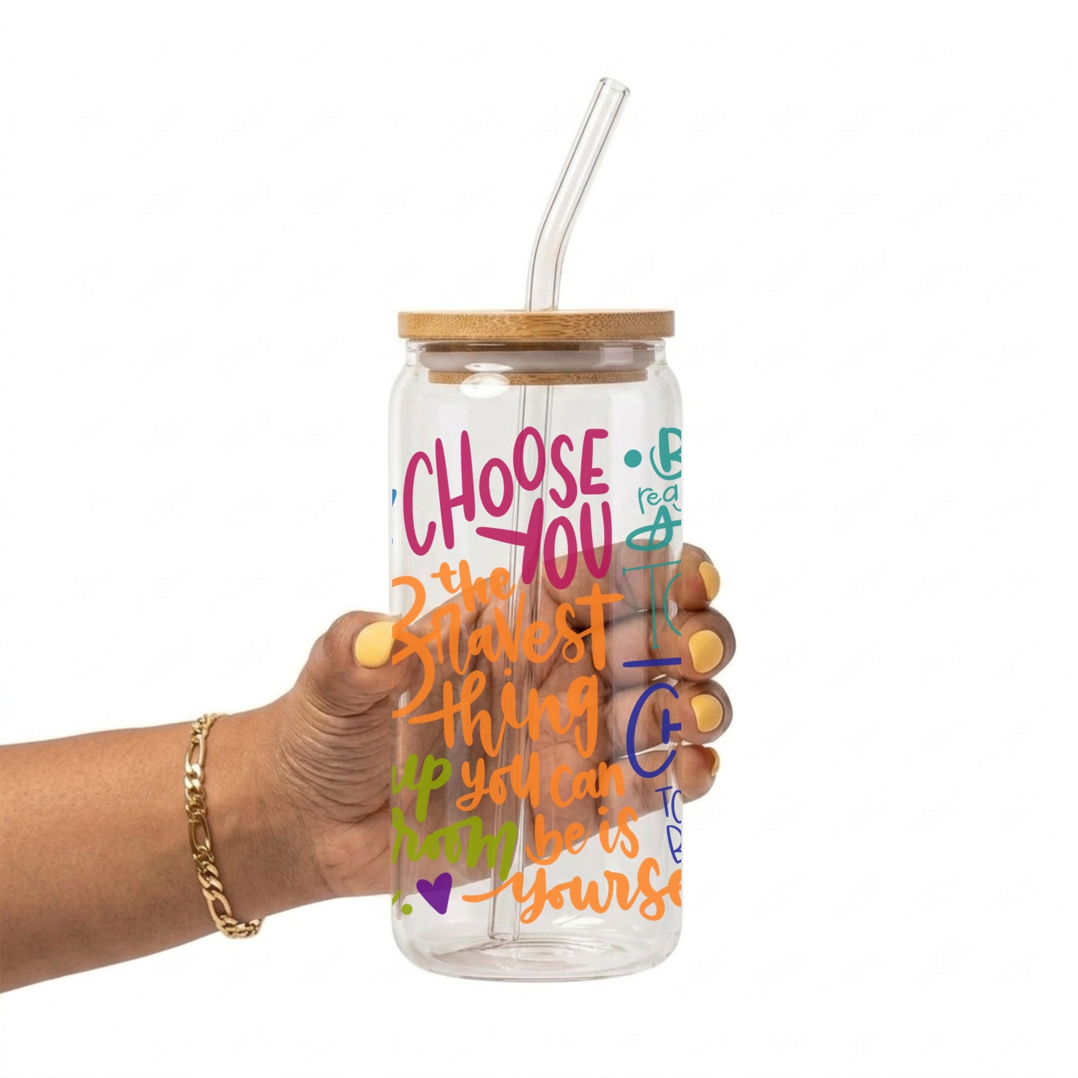 A hand holding a clear glass water bottle with a bamboo lid and clear glass straw. The bottle features colorful hand-lettered text that reads 'CHOOSE YOU 2 the bravest thing you can many be is yourself' in pink, orange, green, and blue.