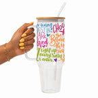A hand with yellow nail polish holds a clear handled tumbler with a bamboo lid and clear straw. The tumbler is printed with colorful inspirational text in blue, pink, orange, green, and purple.