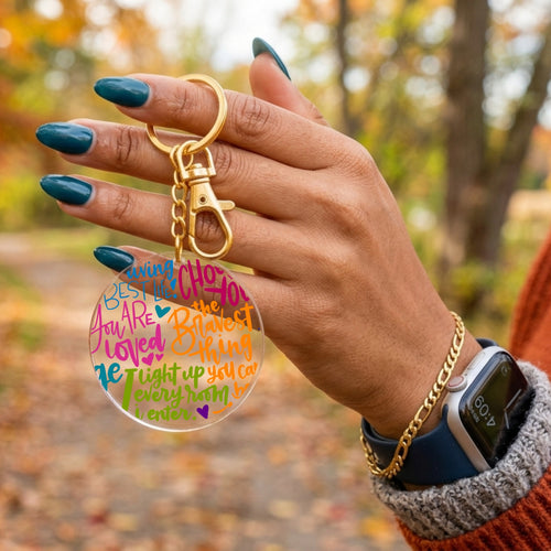A close-up of a hand holding a clear acrylic keychain with colorful inspirational text and a gold-tone key ring and clasp, set against a blurred outdoor autumn background.