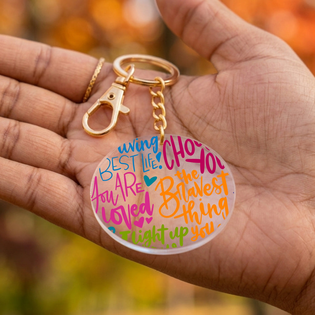 A gold-tone keychain with a clear round acrylic charm printed with colorful motivational hand-lettered text, held in a person's hand against a blurred autumn background.