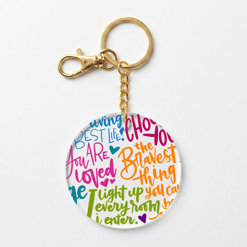 A gold-tone keychain with a round clear acrylic charm featuring colorful hand-lettered text, shown on a clean white background.