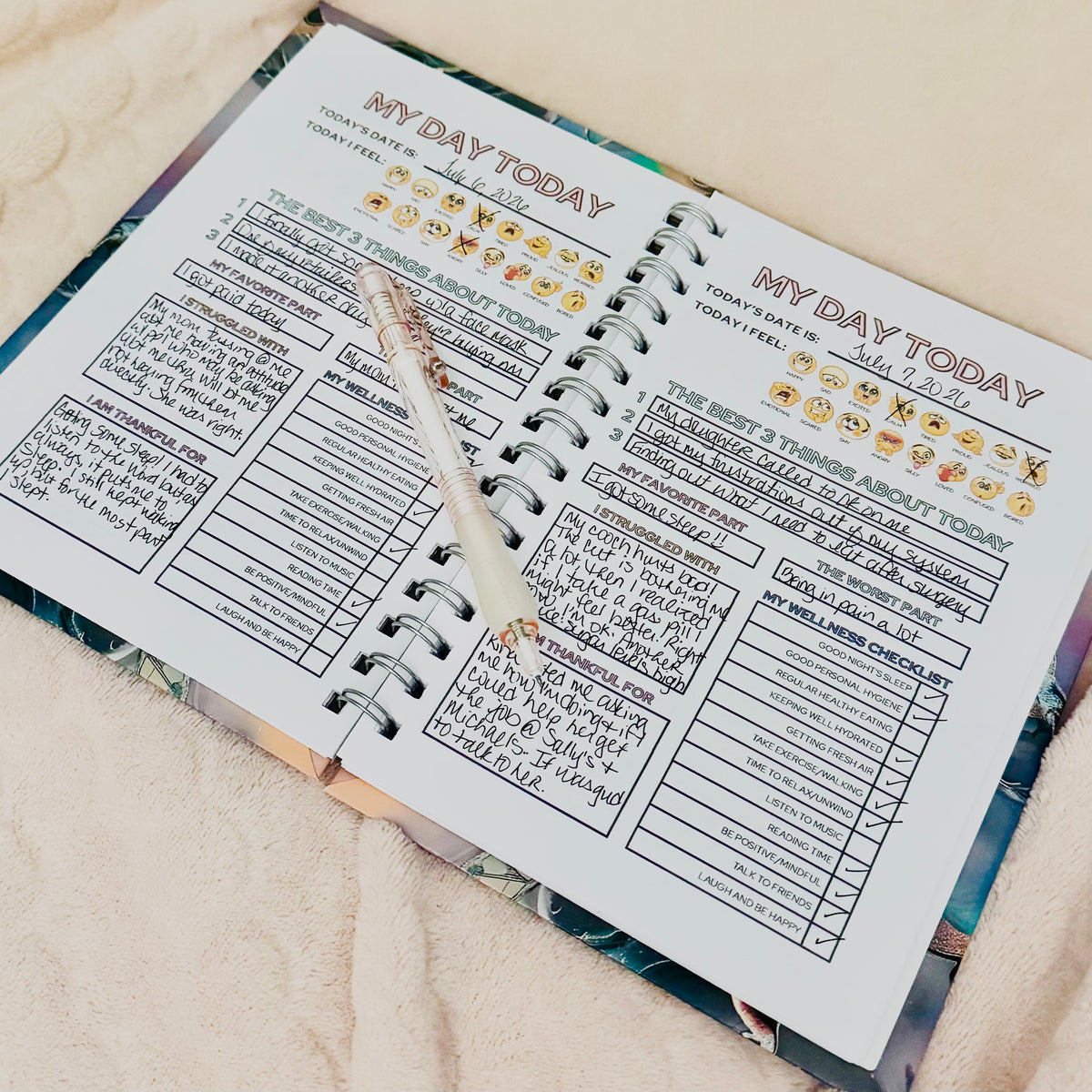 An open self-love journal showing a daily planning page titled 'MY DAY TODAY' with handwritten notes, smiley face checkboxes, and a decorative pen resting across the page, set on a soft beige fabric background.