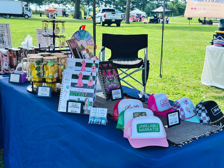 Outdoor market booth display featuring multiple colorful baseball caps, notebooks, and small merchandise items arranged on a blue tablecloth, with a grassy field and event tents in the background.
