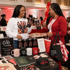 Two women smiling and clinking matching black tumblers at a booth display. The booth features mugs, notebooks, pens, a CoRo & Company sign, and a tote bag that reads "Sisterhood Scholarship Service", with red balloons and a busy event background.