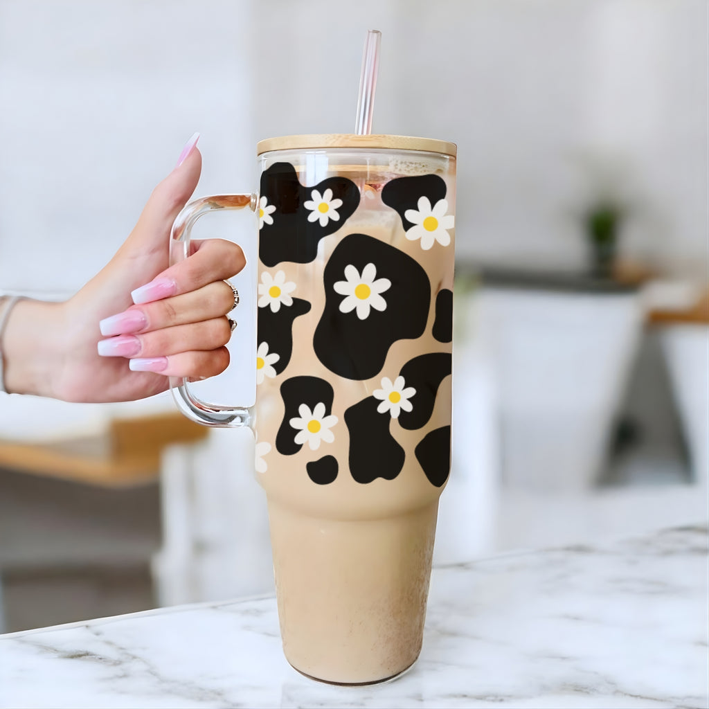 A hand with pink manicured nails holds a tall clear handled tumbler filled with an iced coffee drink. The tumbler has a black and tan cow-print pattern overlaid with small white daisy flowers, and a clear lid with a pink straw.
