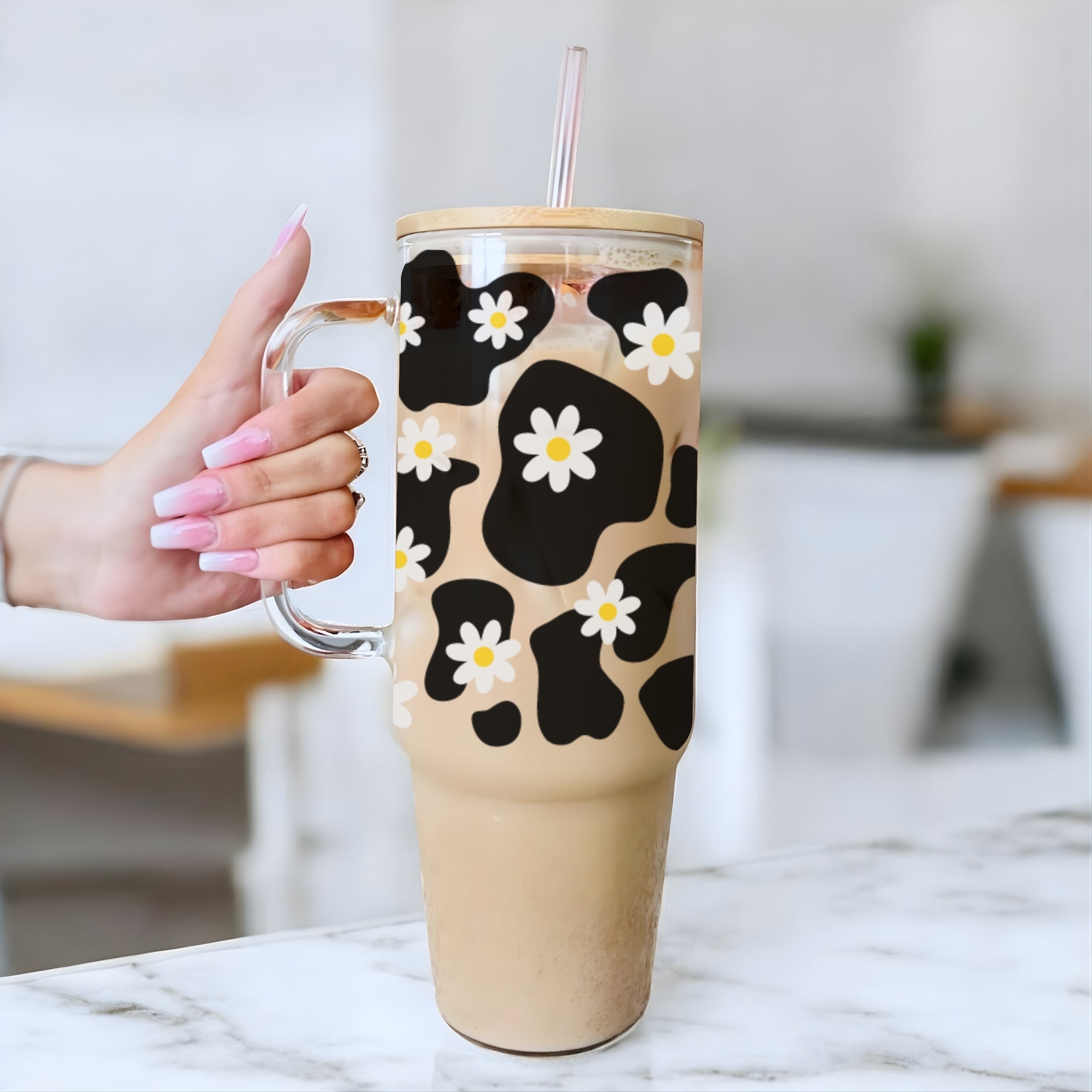 A hand with pink manicured nails holds a tall clear handled tumbler filled with an iced coffee drink. The tumbler has a black and tan cow-print pattern overlaid with small white daisy flowers, and a clear lid with a pink straw.