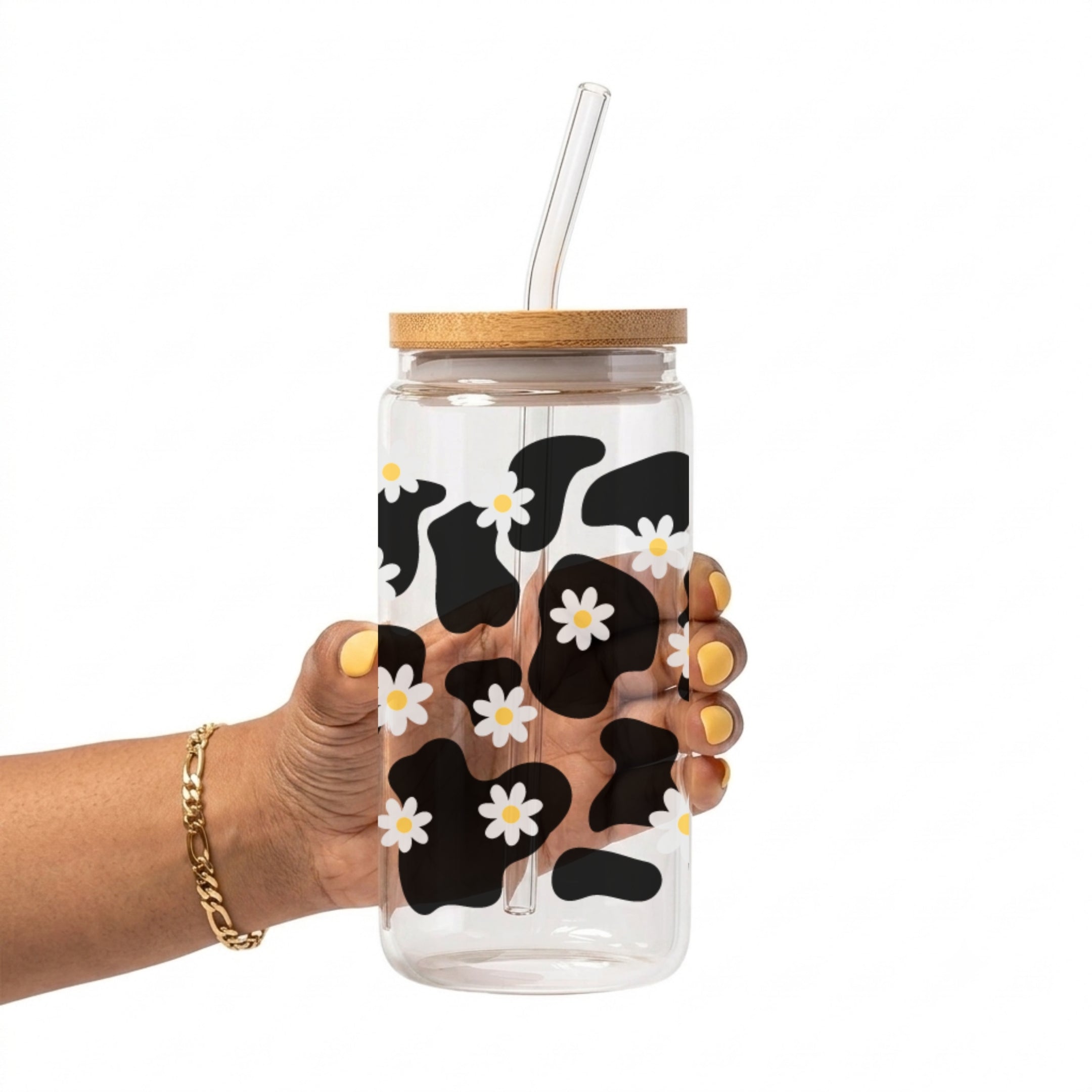 A hand holding a clear glass tumbler with a bamboo lid and clear glass straw, decorated with a black and white cow print pattern and small white daisies with yellow centers.