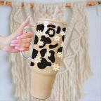A hand with pink manicured nails holds a clear handled tumbler filled with a light brown iced coffee or latte. The tumbler has a black and beige cow-print style pattern overlaid with white daisy flowers, and a clear straw.