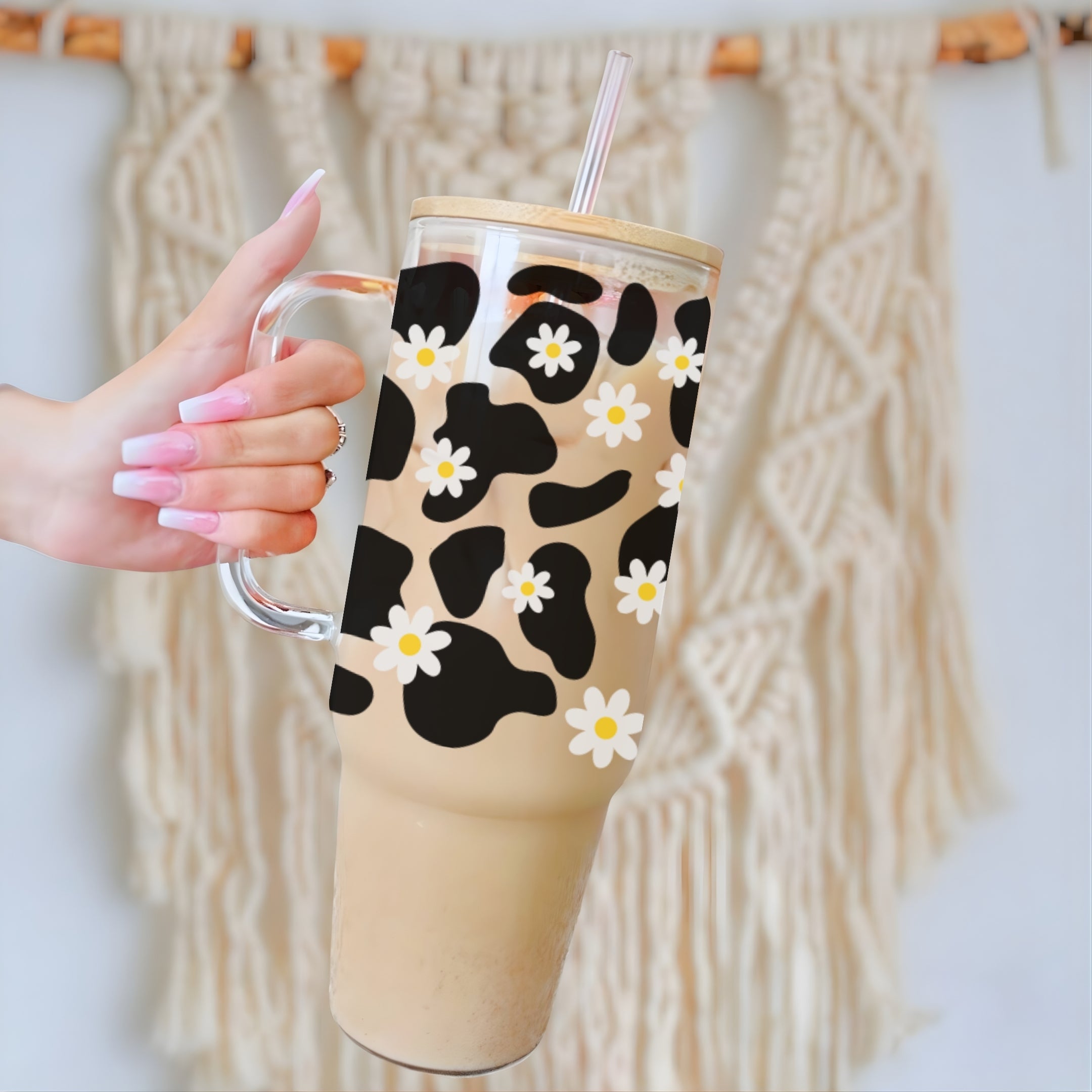 A hand with pink manicured nails holds a clear handled tumbler filled with a light brown iced coffee or latte. The tumbler has a black and beige cow-print style pattern overlaid with white daisy flowers, and a clear straw.