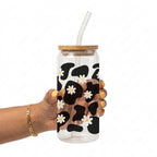 A hand holding a clear glass tumbler with a bamboo lid and clear straw, decorated with a black and white cow print pattern and small white daisies with yellow centers.