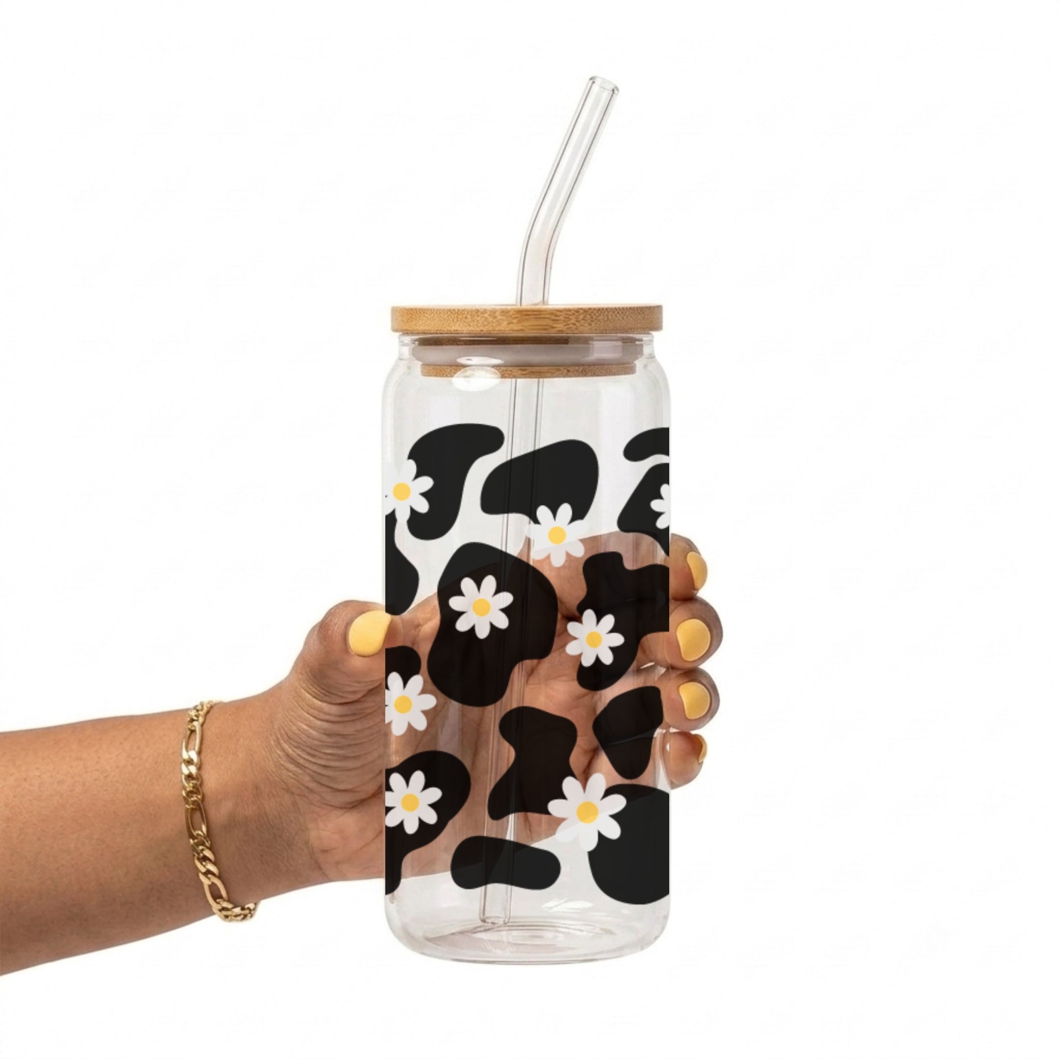 A hand holding a clear glass tumbler with a bamboo lid and clear straw, decorated with a black and white cow print pattern and small white daisies with yellow centers.