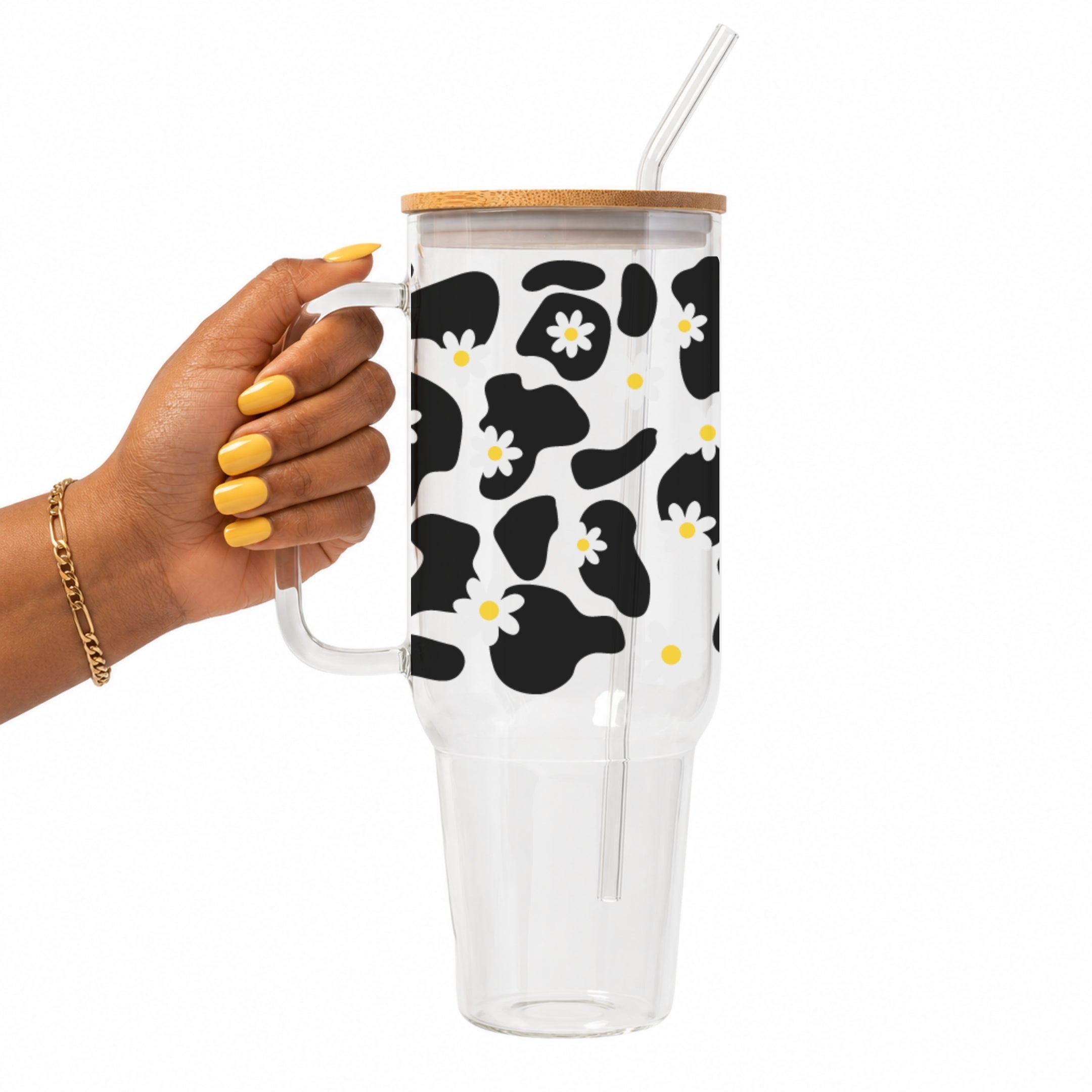 A hand with yellow nails holds a clear handled tumbler with a bamboo lid and clear straw. The tumbler has a white background with a black cow print and small white daisy flowers with yellow centers.