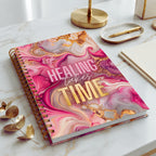 A spiral-bound self-love journal with a pink, purple, and gold marble-patterned cover lies on a white marble desk. The cover features the text 'HEALING TAKES TIME' in pink and gold lettering.