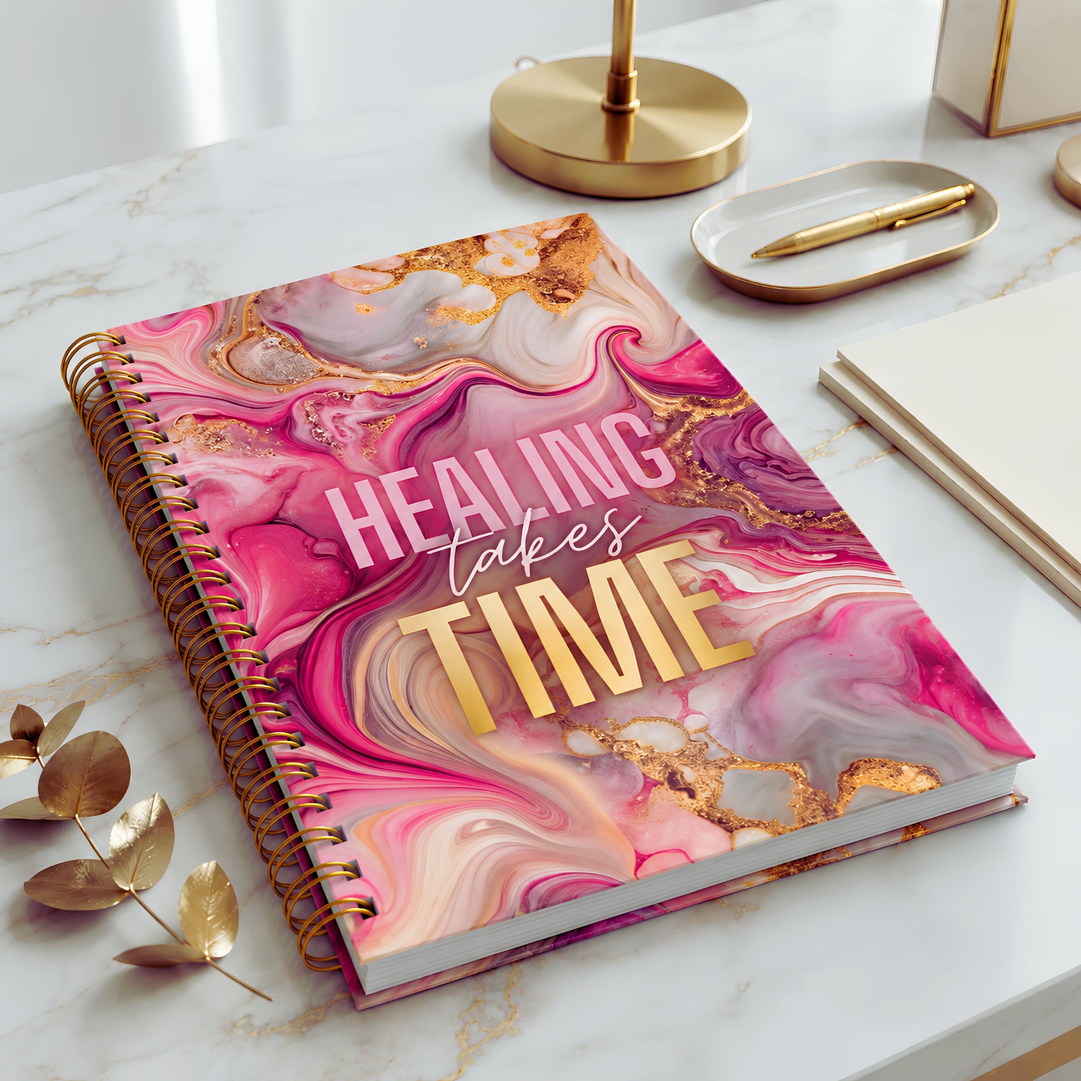A spiral-bound self-love journal with a pink, purple, and gold marble-patterned cover lies on a white marble desk. The cover features the text 'HEALING TAKES TIME' in pink and gold lettering.