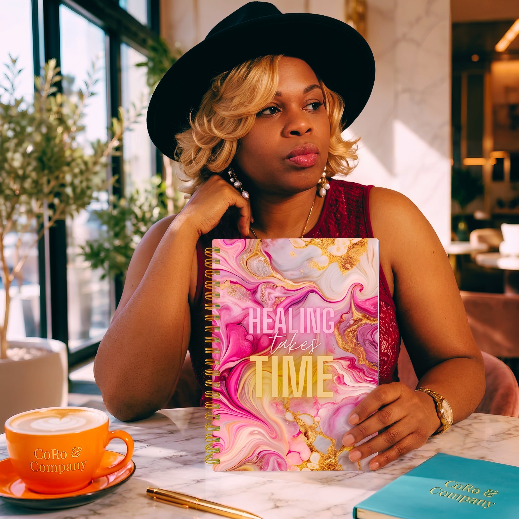 Nik Austin wearing a black hat sits at a cafe table holding her handmade pink and gold marbled spiral notebook that reads 'HEALING TAKES TIME'. An orange CoRo & Company coffee cup is on the table next to her, with a matching teal CoRo & Company notebook in the foreground.